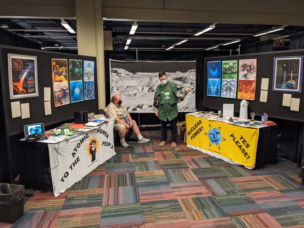 Photo of the "Man and Atom" display at Chicon 8. There are two tables with exhibits on them, and hortatory table fronts. Above these are displays of art. In the background is the famous Lunar Orbiter low-angle photo of Copernicus, printed on cloth.