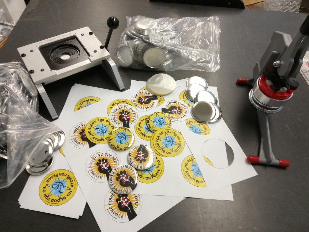 Badge parts, finished badges, badge press, and paper cutter, laid out on a worktable. Two designs promoting civil atomic energy are in evidence, with slogans "Atomic Power to the People!" and "No Blood for Oil ― Atoms for Peace".