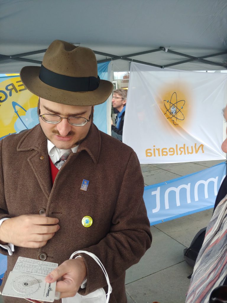 A man (publius) in an overcoat and fedora, showing off a circular slide rule. Pro-nuclear-energy protesters and signs in the background.