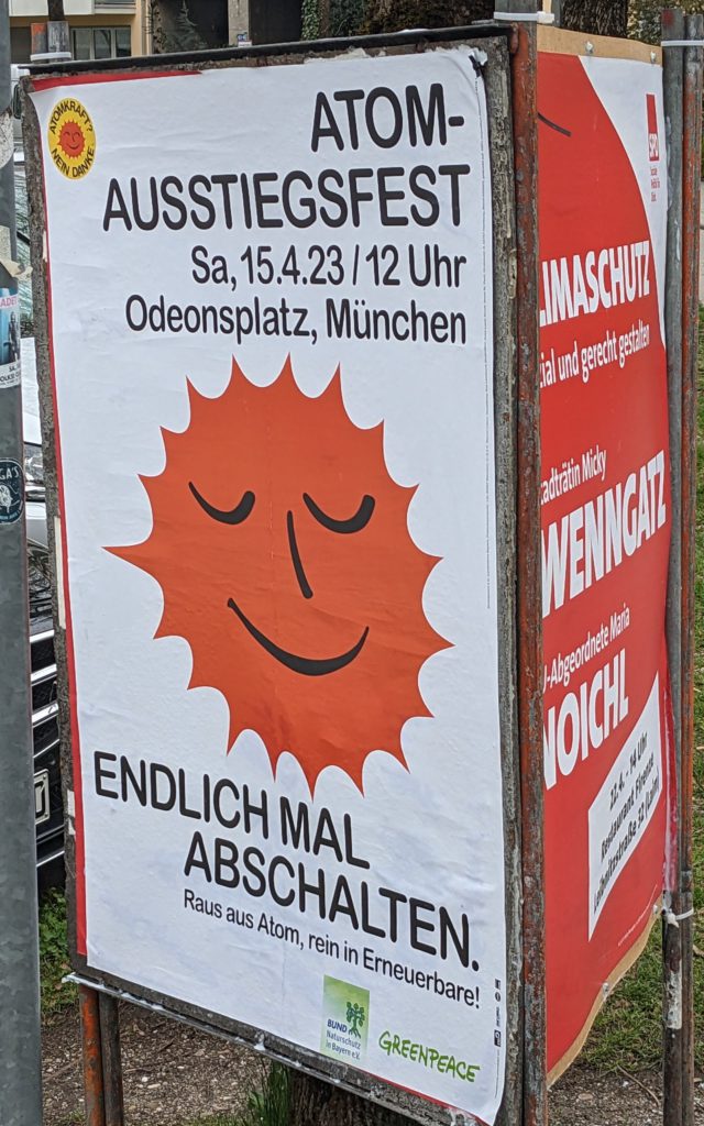 A typical scene in Munich, political posters set up in a public place. The one facing the camera is advertising a celebration of the shutdown of the last nuclear power plants in Germany. The one adjacent to it is advertising a talk about the alleged climate protection policy of the governing coalition.