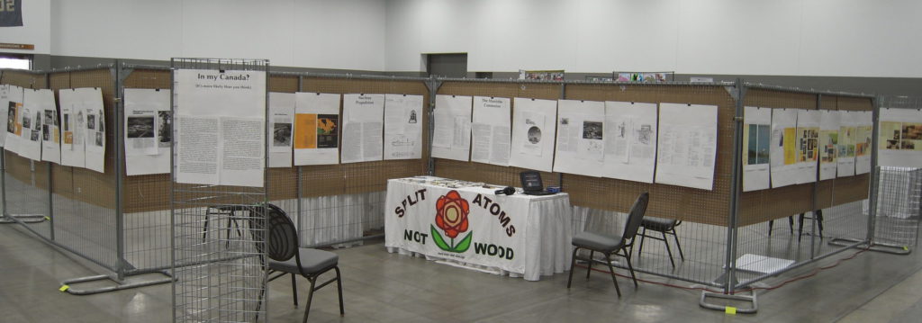 A view of the "Man and Atom" display at Pemmi-con in Winnipeg, Manitoba. A table can be seen draped with the "atomic flower" logo and the motto "Split Atoms Not Wood". On it there is a portable video player. Along four sections of fence panelling, and on freestanding grid constructions, are posterboards with panels covered with text and images, including many pages scanned from public-information documents.
