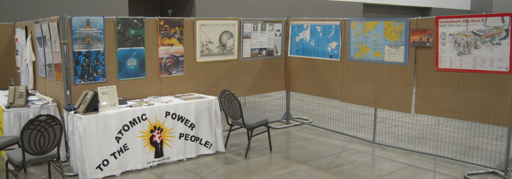 A view of part of the "Man and Atom" display at Pemmi-con in Winnipeg, Manitoba. Two tables can be seen, one facing the viewer and draped with the "radiant atom clutched in a fist" logo and the slogan "Atomic Power to the People". A variety of nuclear-related posters and other materials is displayed along sections of fencing covered with pegboard.