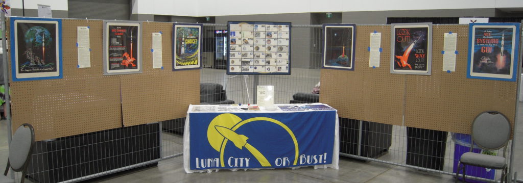 Luna Project display at Pemmi-con in Winnipeg, Manitoba. In the centre of the image a display of postal covers hangs over a table draped with a cloth displaying the blue-and-yellow Luna Project "rocket" logo at front. Along the panels to both sides are hung a set of six prints by Frank Kelly Freas.