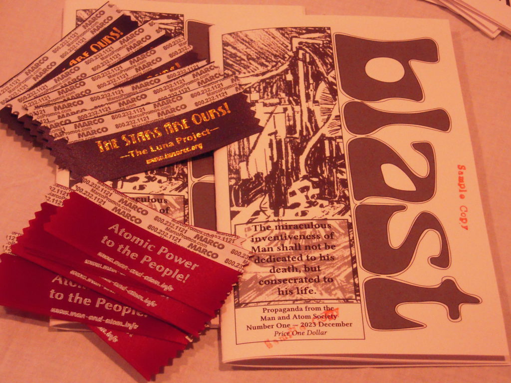 Two copies of a booklet with the title "blast", stamped "Sample Copy" in red, a pile of red badge-sticker ribbons with the slogan "Atomic Power to the People" in silver, and a pile of dark blue ribbons with "The Stars Are Ours!" in gold. White balance is seriously off, giving the whole image a pink tint.