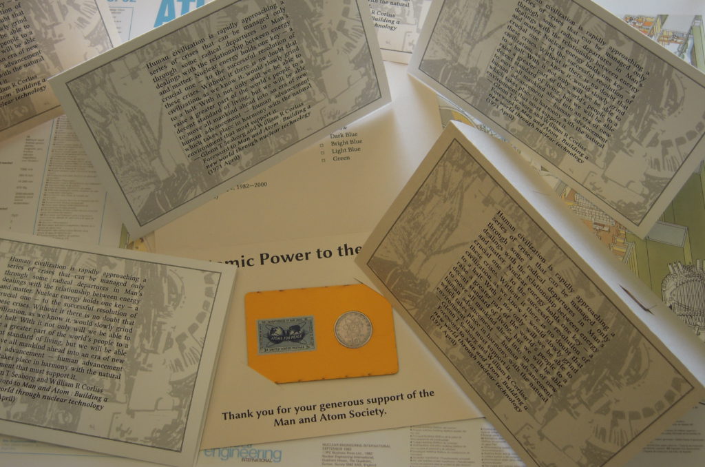 Several folded letter-size sheets of cardstock, printed with text and images. One lies open, revealing a bright yellow card perforated with a hole, in which a coin is pressed. Affixed to the card is a blue "Atoms for Peace" postage stamp, 