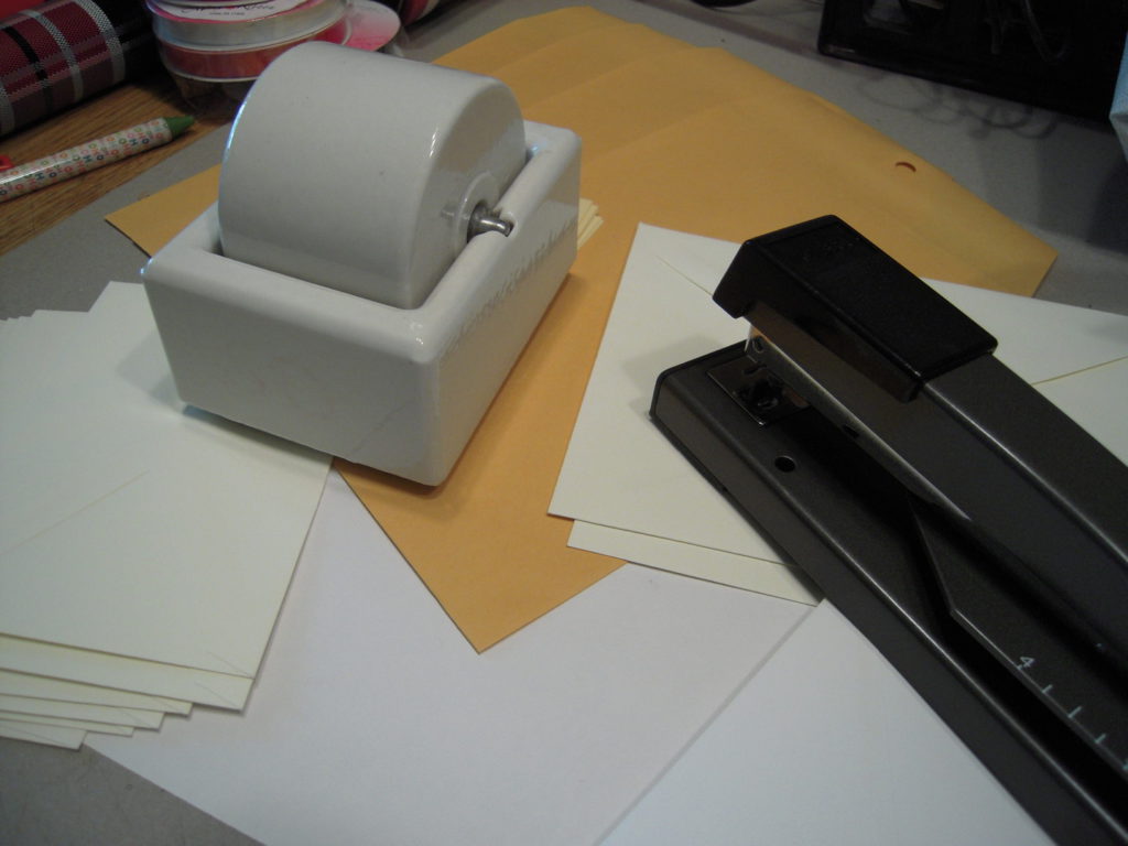 On a worktable, a white ceramic roller-moistener sits, accompanied by envelopes in manila, cream, and white, and the working end of a long-arm stapler