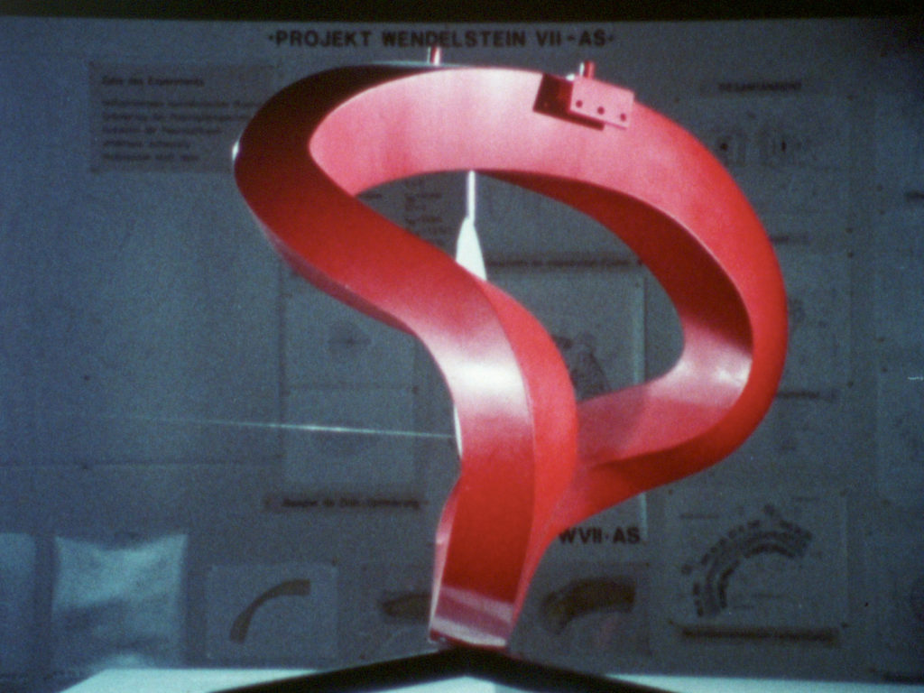 A strange red shape, square in cross-section and forming a twisted loop, against an indistinct background of diagrams