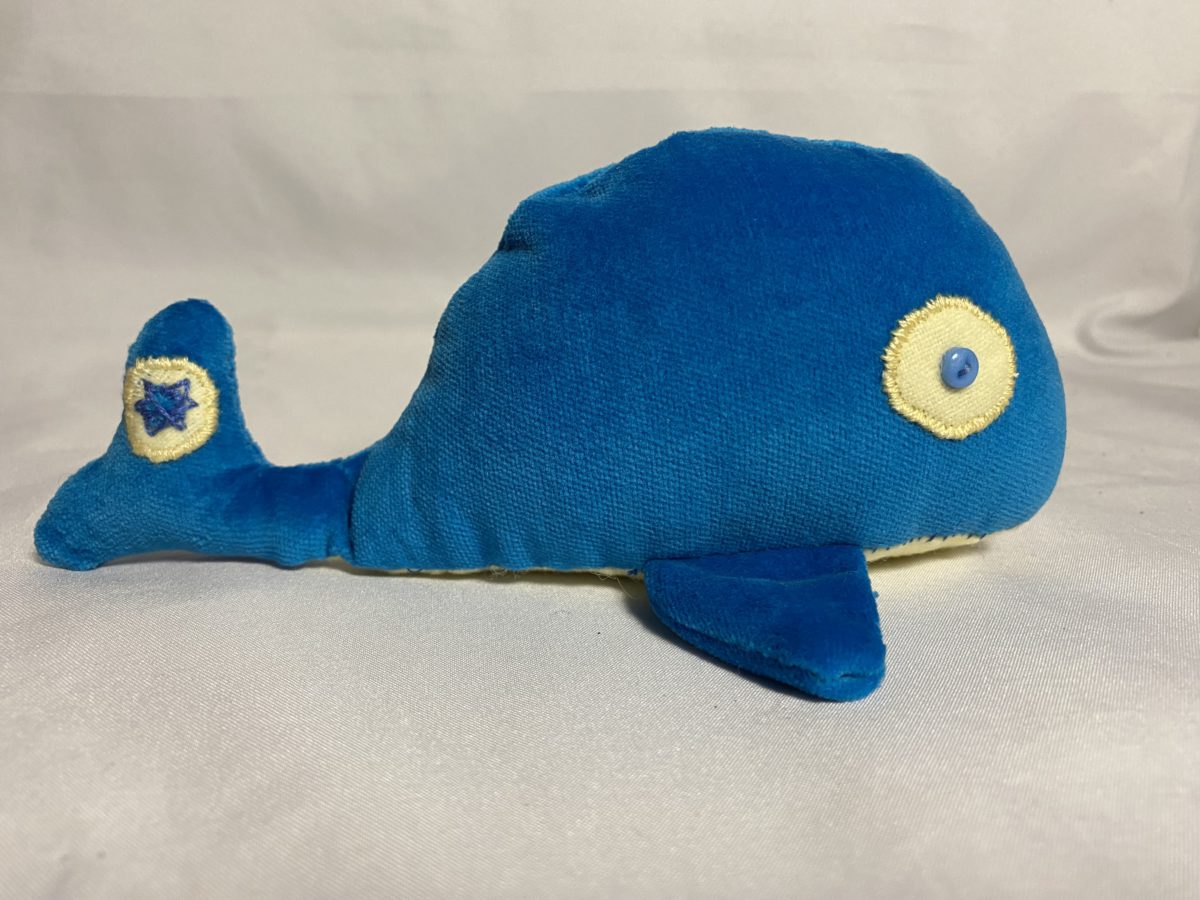 Blue and yellow plushie whale with a "Nuclear Power Yes Please" badge on the tail