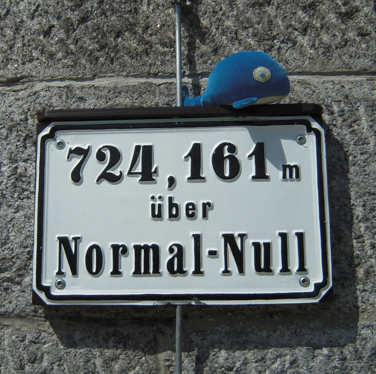 A blue plushie resembling a sperm whale, sitting atop a sign proclaiming the elevation to be 724 meters above Normal-Null